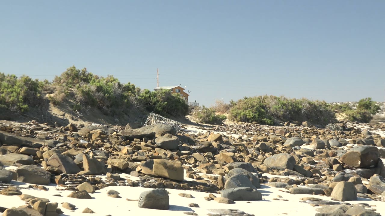 La Bocana beach frame showing scattered rocks over bright sand, scrubby vegetation on the left, ocean water beginning to show on the right, and no visible people.