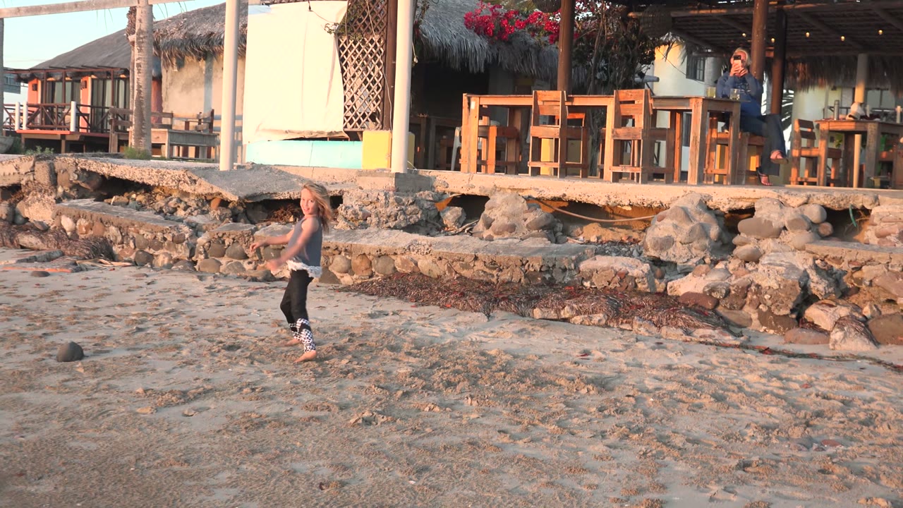 In San Carlos, Mexico, a child runs across the sandy beach near a stone wall, with a thatched-roof shelter, wooden tables, palms, and an adult visible behind.