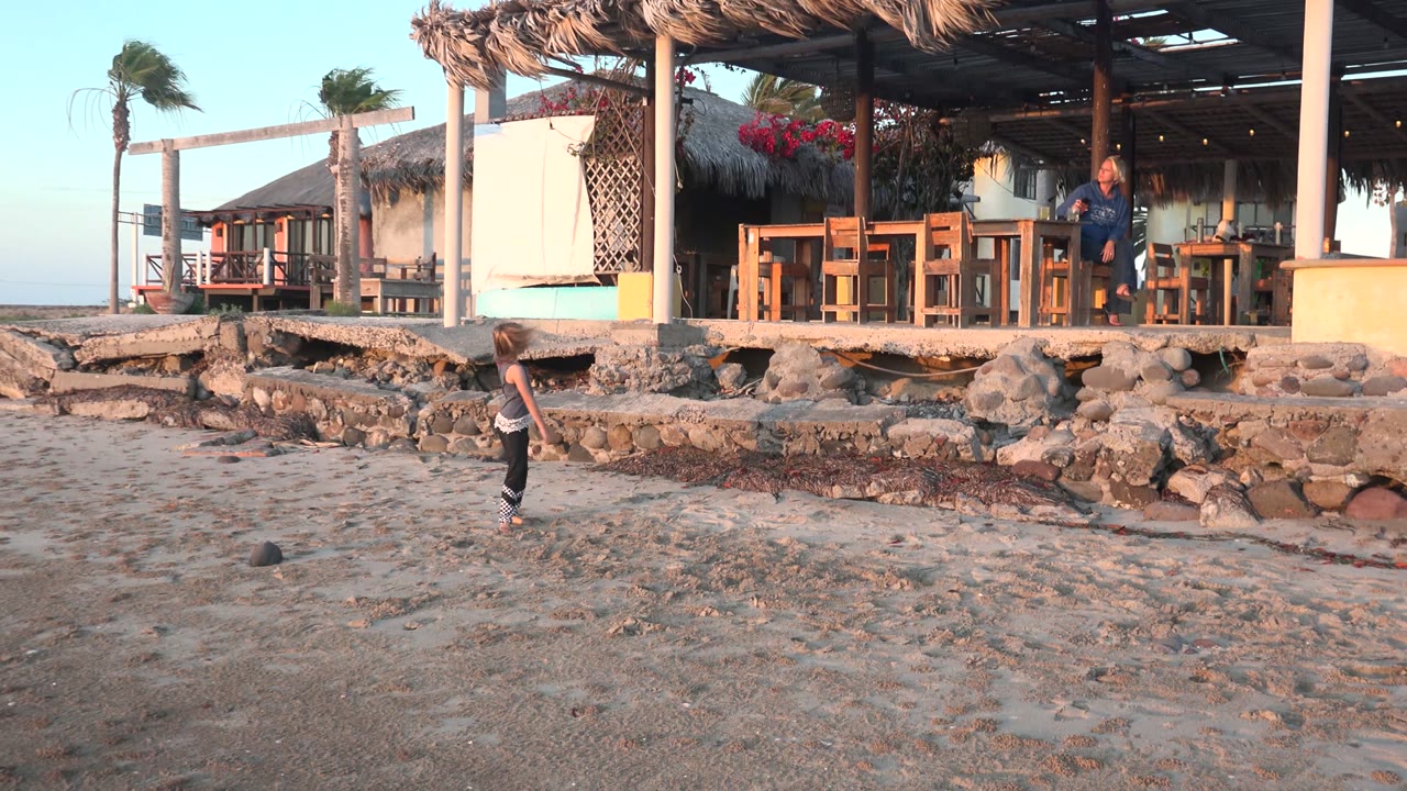 In San Carlos, Mexico, the child runs across the open sand while the thatched shelter, stone wall, wooden furniture, palms, and adult remain in the background.
