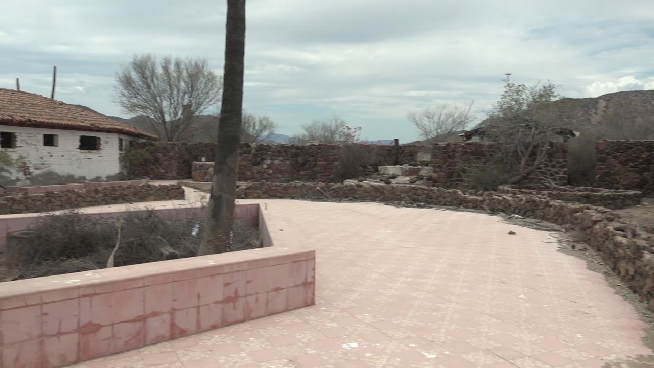 At the start of the Mulege, Baja California Sur clip, a pink-tiled courtyard, palm trunk, stone walls, dry plants, and a white building with a red tile roof appear under overcast sky with no visible people.