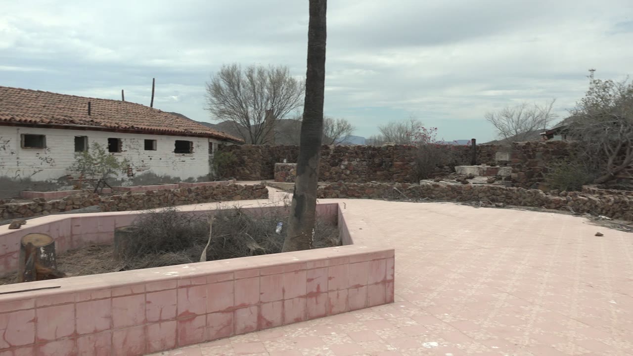 At 0:02 in Mulege, Baja California Sur, the palm trunk sits near center over the pink courtyard wall while stonework and the white building with small dark windows remain in view, with no visible people.