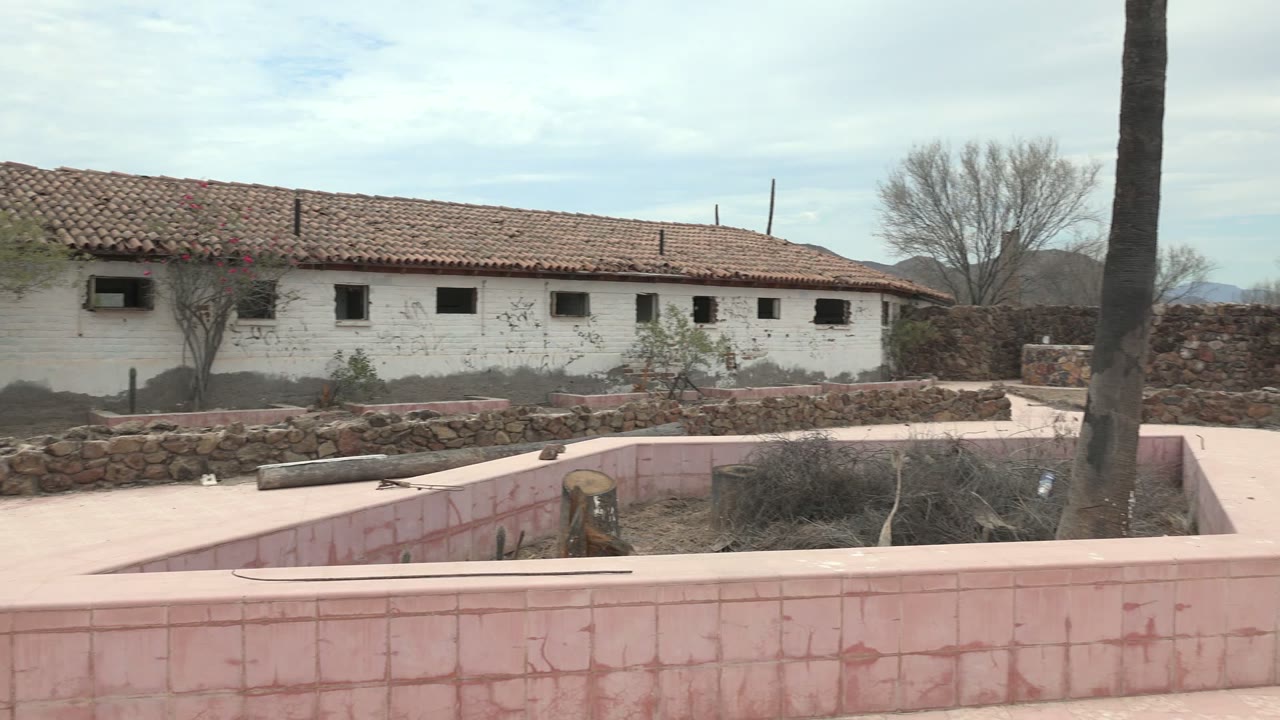In Mulege, Baja California Sur, a white building with a red tile roof stands behind pink courtyard walls and dry landscaping, with a small person visible on the roof near the center.