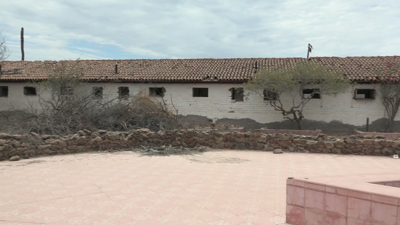 At 0:00 in Mulege, Baja California Sur, a white tiled-roof building runs across the background behind dry bushes, a low stone wall, and a pink concrete courtyard, with no visible people.