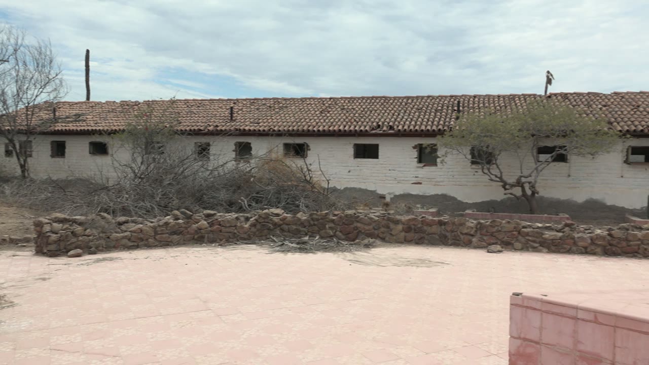 Dry courtyard with white tiled-roof building, stone wall, and brush.