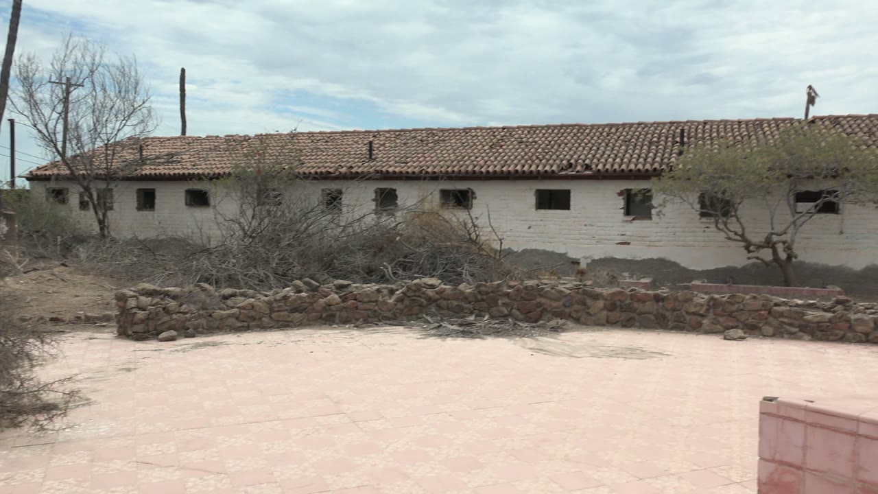 At 0:02 in Mulege, dry shrubs and bare trees sit between the pink courtyard and the long white building, with the low stone wall crossing the frame and no visible people.