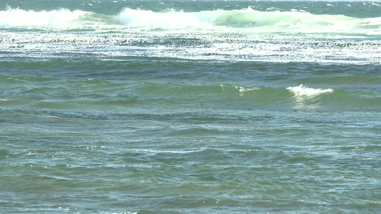 At La Bocana, the frame holds textured blue-green water in the foreground while distant waves break into white foam under a pale sky, with no visible people.
