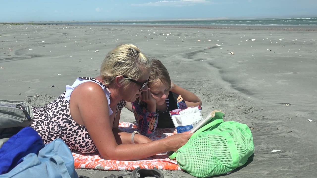 Woman and child look down at reading material on a sandy beach.