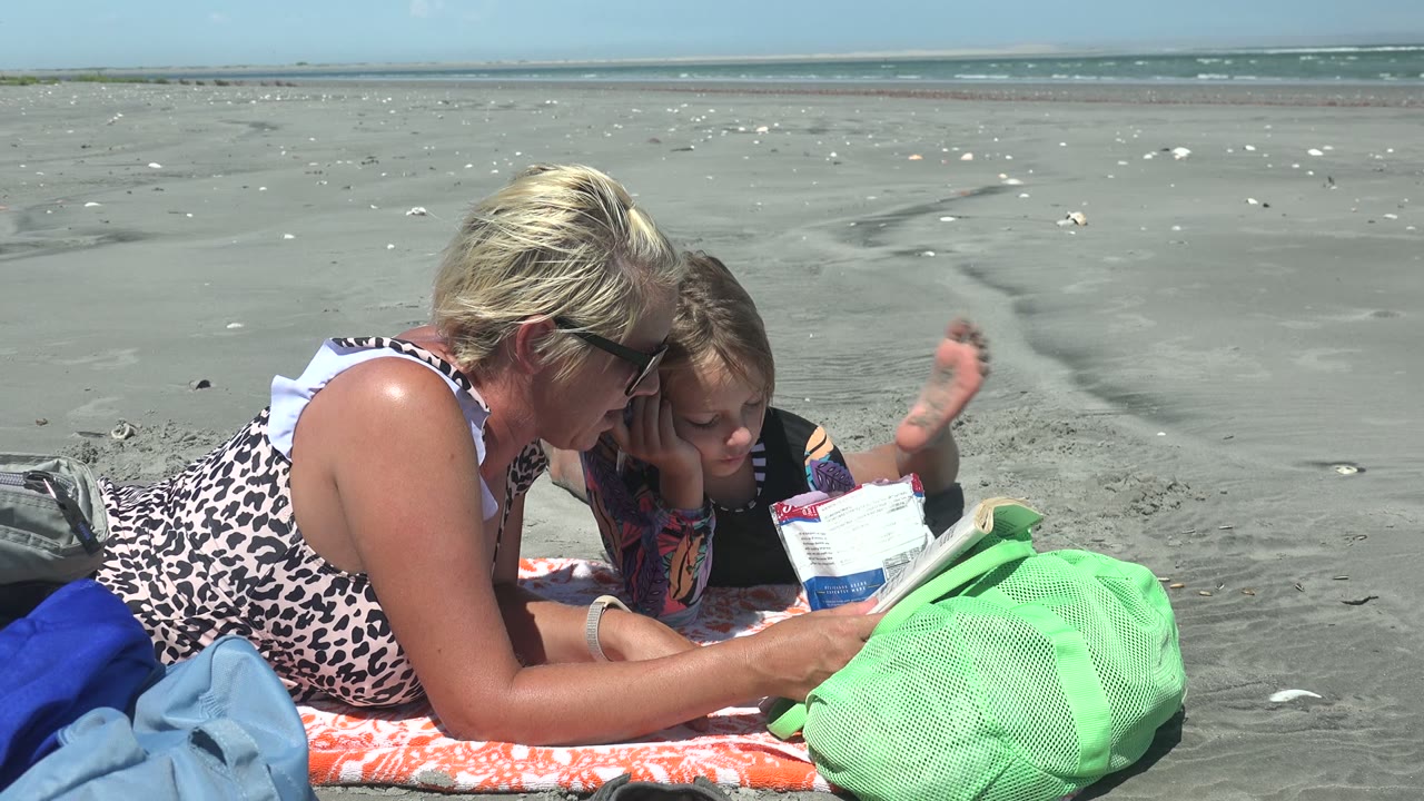 In La Bocana, Mexico, the child raises an arm while lying beside the woman, with reading material between them and open sand stretching to the ocean.