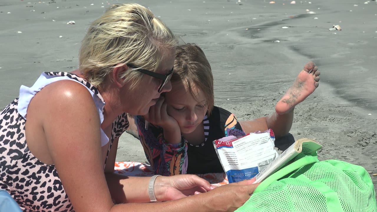 At 0:00 in La Bocana, Mexico, a woman in sunglasses leans close to a child as they look down at reading material on the sandy beach beside a green mesh bag.