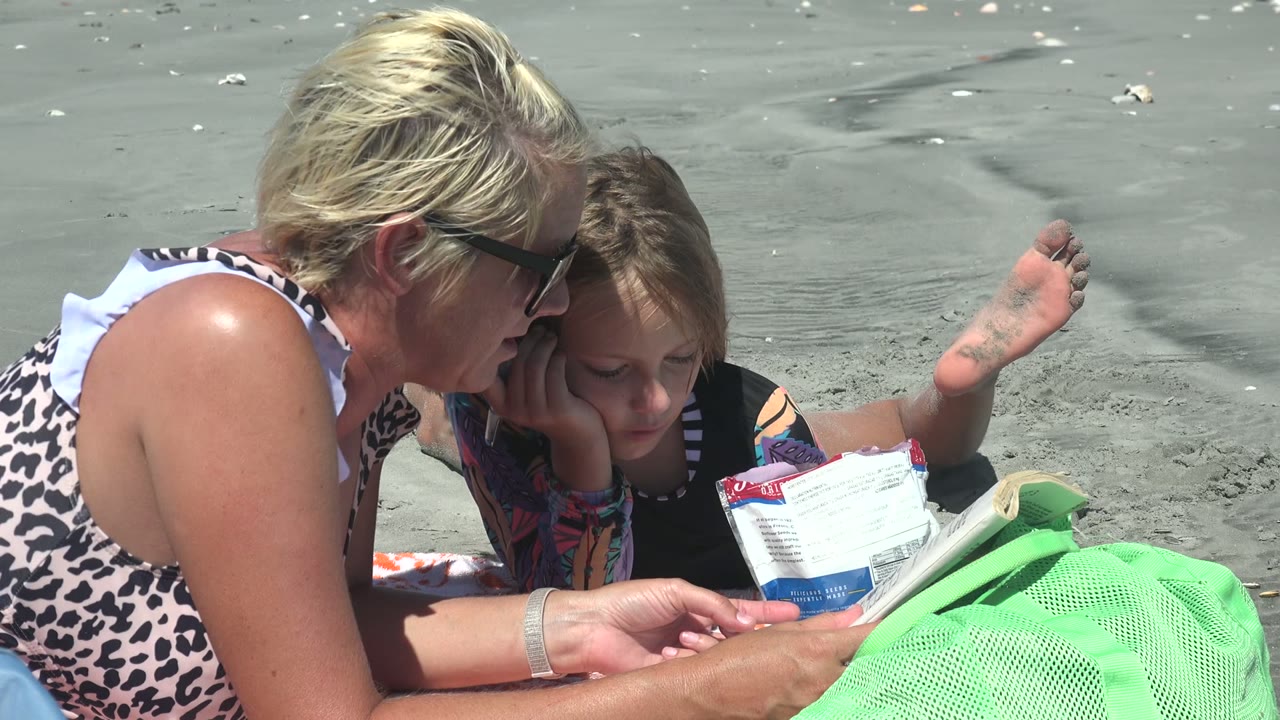 Woman and child remain close together on the sand with reading material in front of them.