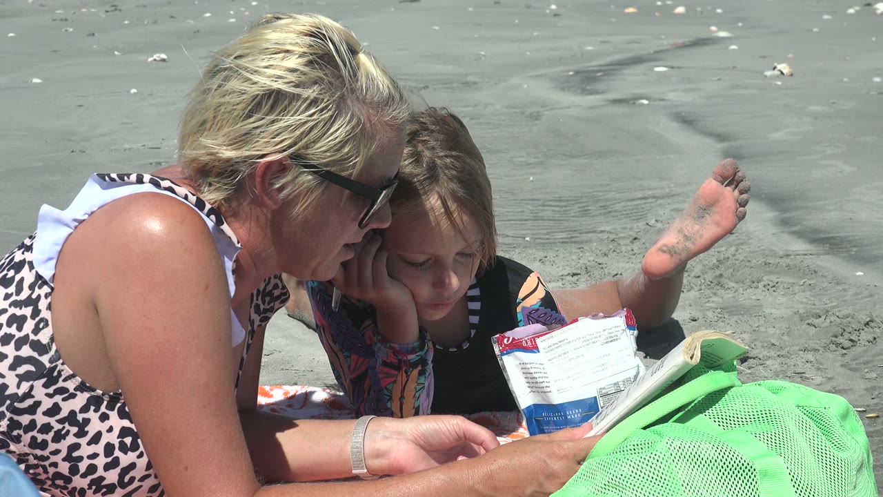 At 0:02 in La Bocana, Mexico, the child rests close to the woman while a small piece of reading material and the bright green mesh bag anchor the lower foreground.