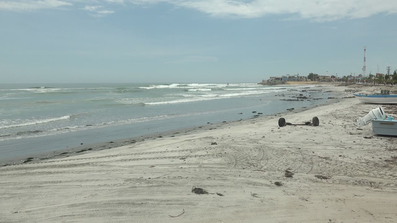 Punta Abreojos beach view with pale tire-marked sand in the foreground, small shore boats at right, white waves offshore, distant coastal buildings, and no visible people.