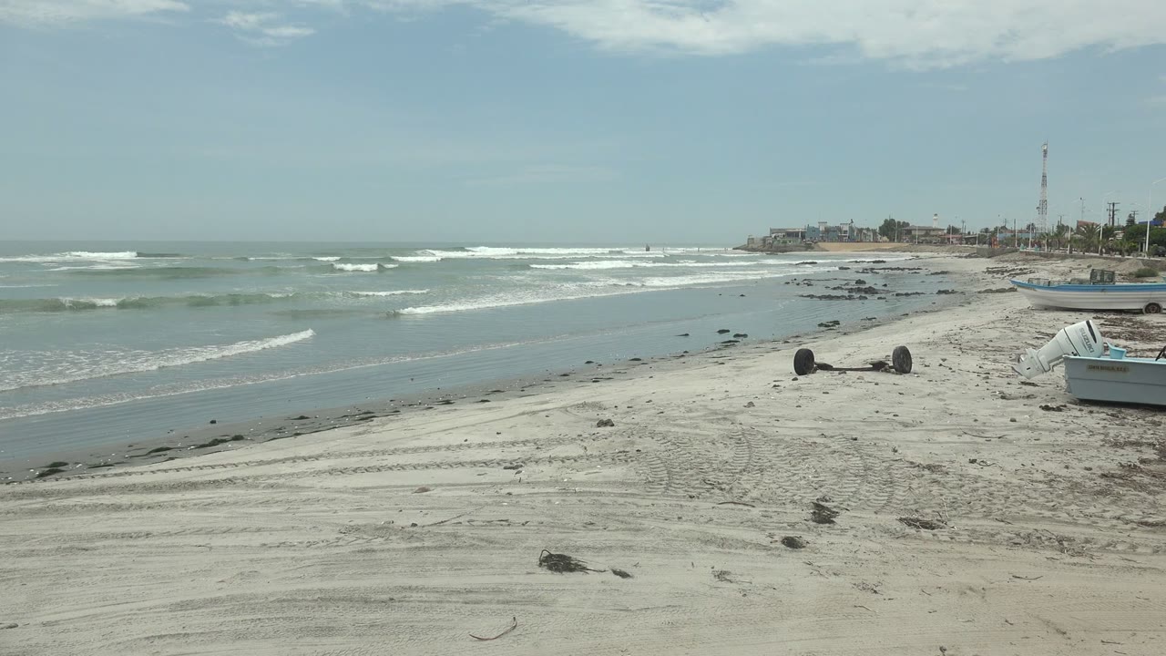 Punta Abreojos beach frame with low waves breaking in bands, tire-marked sand and dark rocks in the foreground, a small shore boat at right, distant buildings, and no visible people.