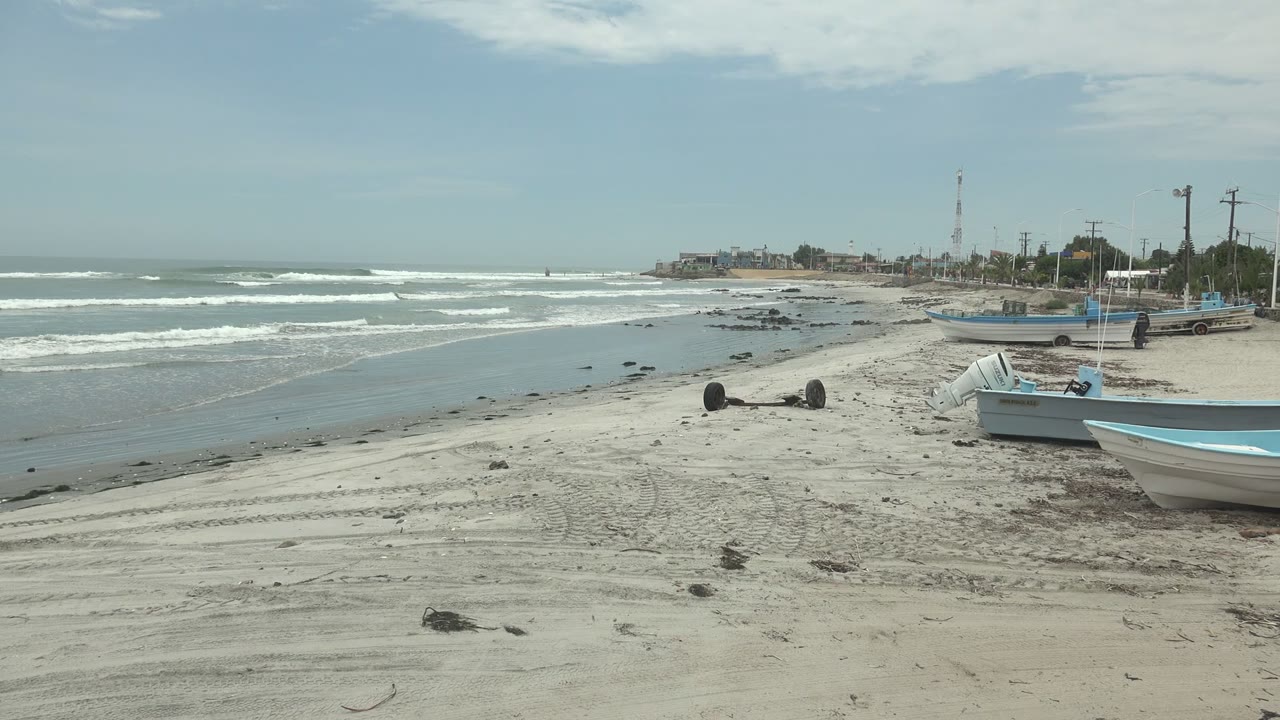 Wide view of Punta Abreojos beach with small blue and white boats on pale sand, ocean waves at left, distant buildings and a communication tower on the horizon, and no visible people.