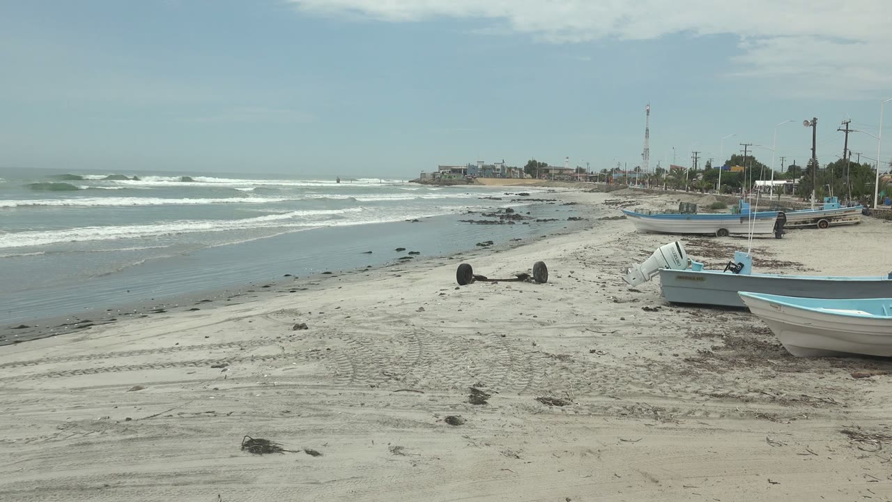 Small blue and white boats rest on the sandy Punta Abreojos shore, with foamy waves behind them, scattered dark rocks or debris near the waterline, and no visible people.