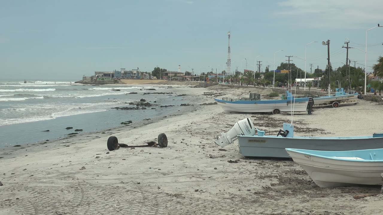 Wide view of fishing boats on sand at Punta Abreojos, Baja California Sur, with gentle surf at left, shoreline buildings and a communication tower in the background, and no visible people.