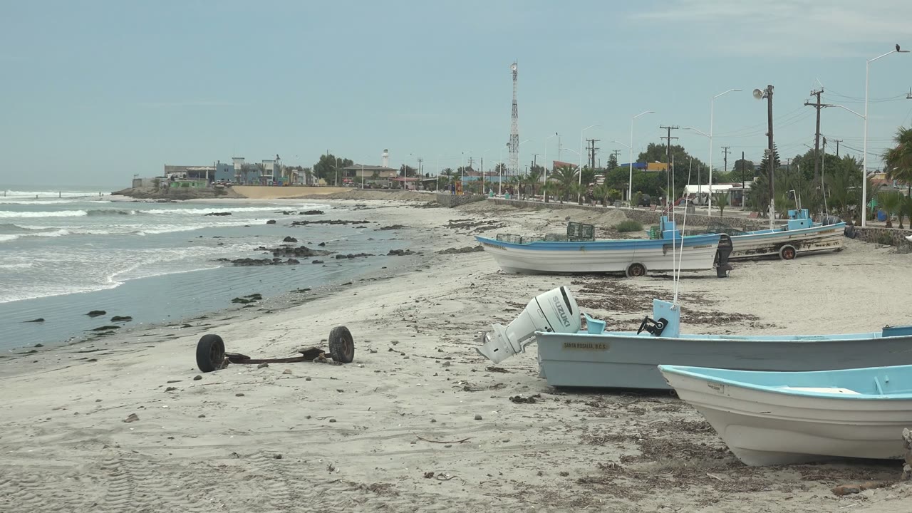 Blue and white boats on sand near waves and shoreline buildings.