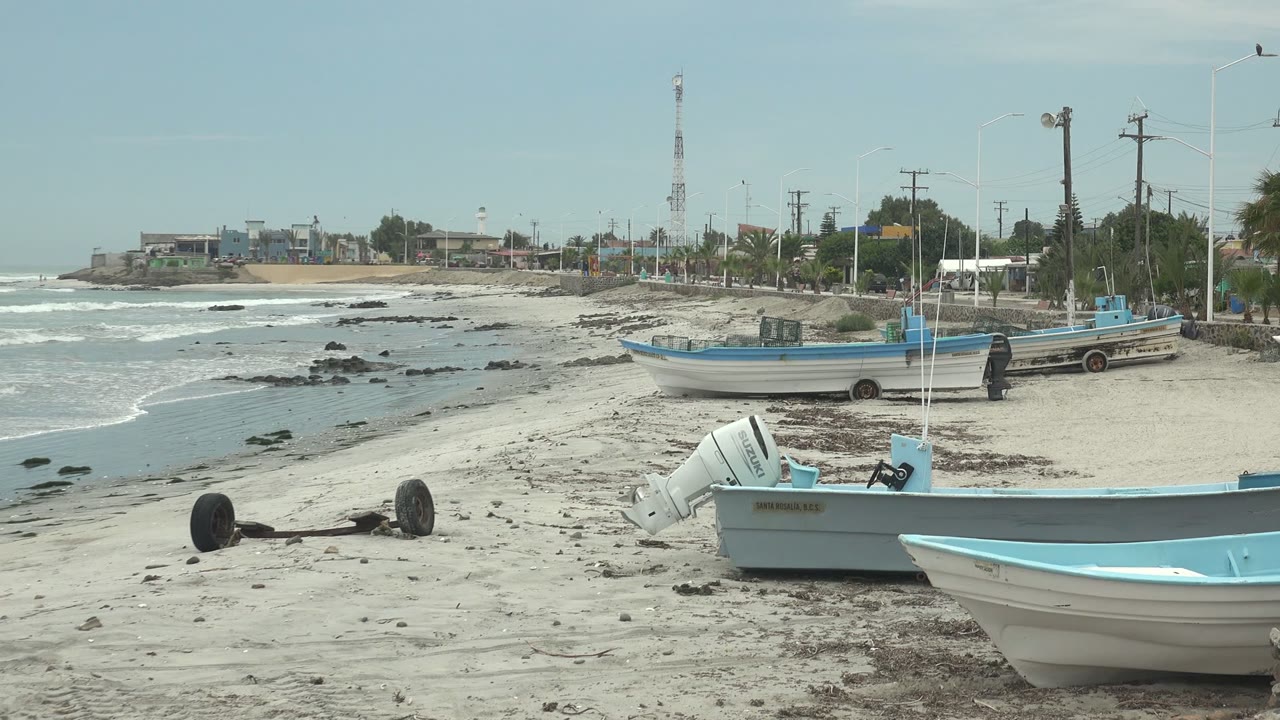 A low beach view in Punta Abreojos, Baja California Sur shows several blue-trimmed fishing boats on sand, surf lines along the left side, roadside poles behind them, and no visible people.