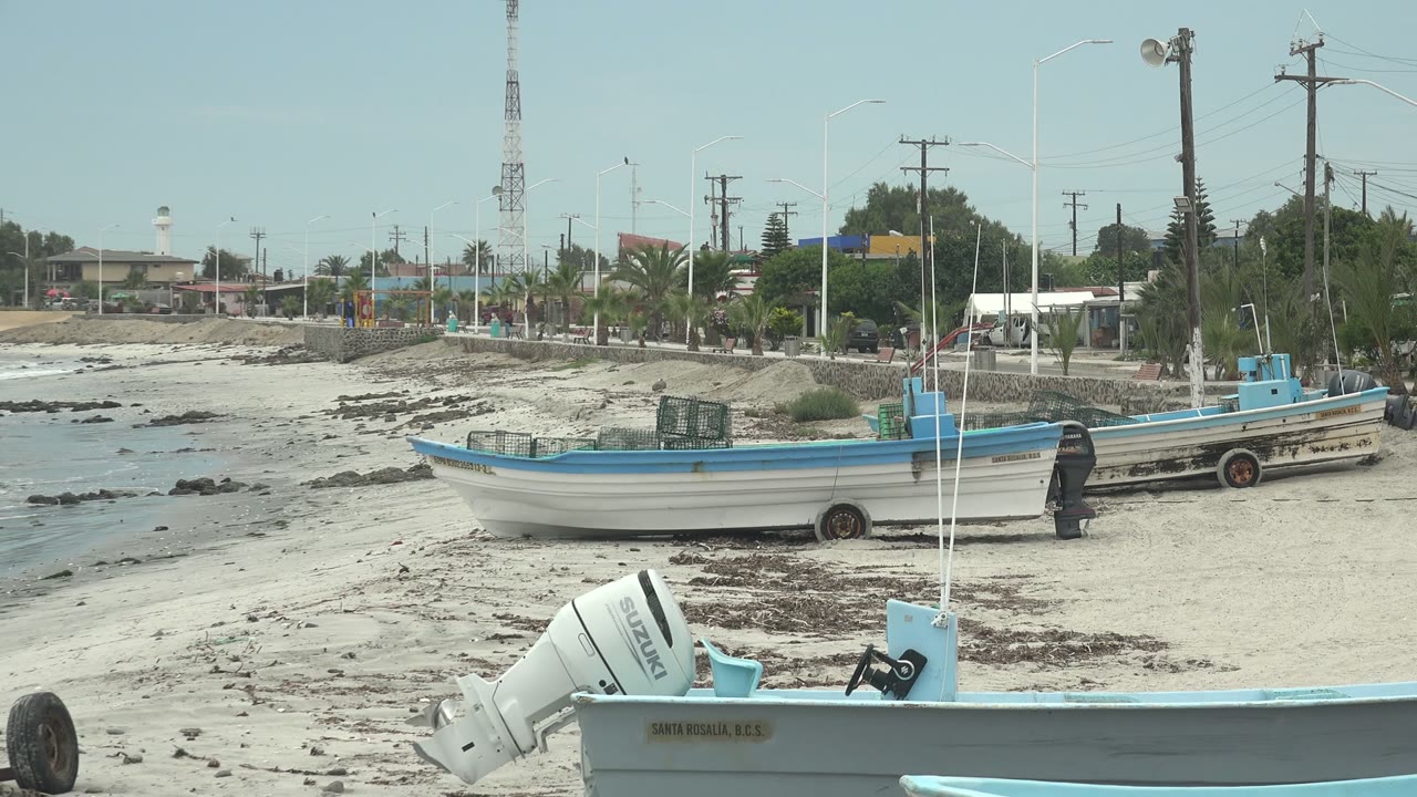 At 0:00 in Punta Abreojos, Baja California Sur, blue and white boats rest on pale sand with seaweed patches, a low seawall, palms, buildings, and distant people in the background.