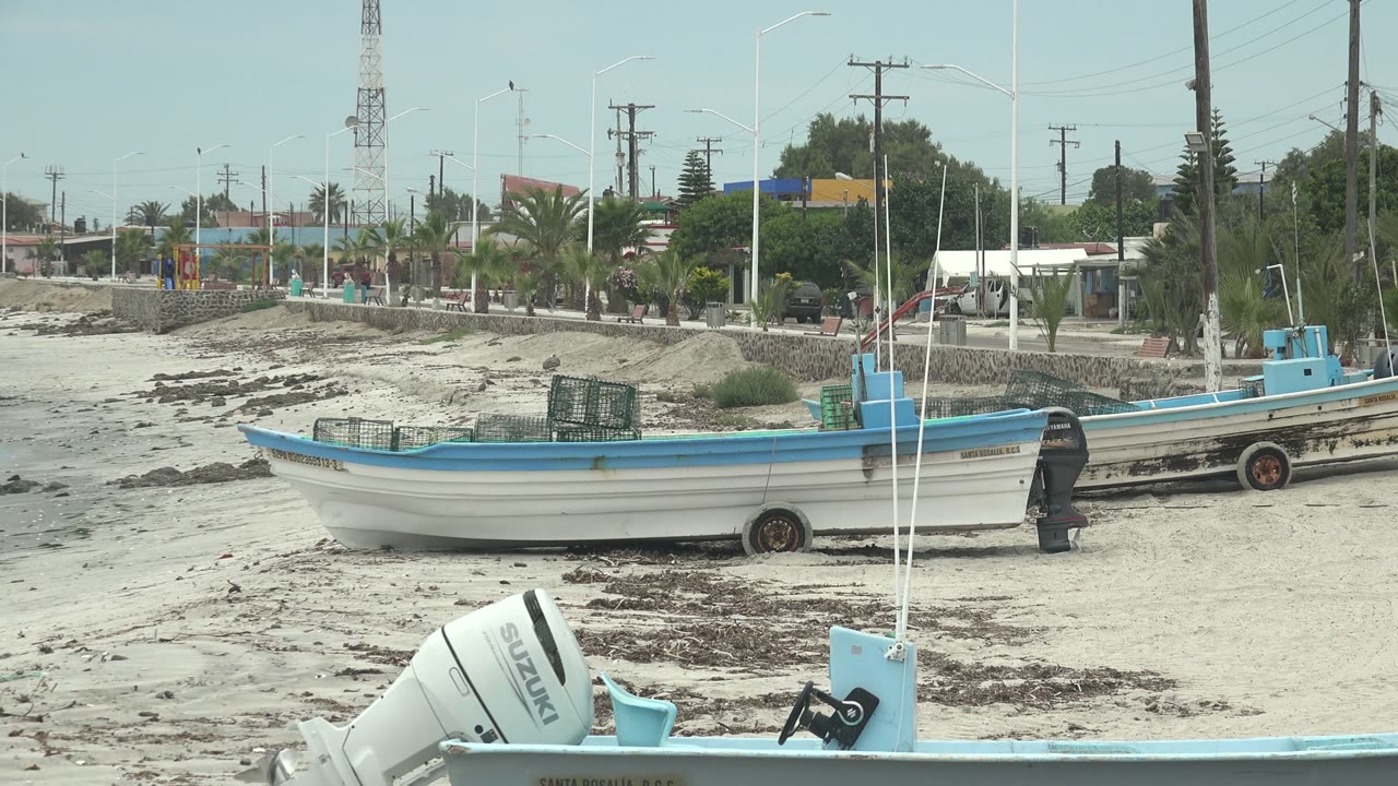 At 0:02 in Punta Abreojos, the midground boat stretches across the sand with crates aboard, another boat behind it, and the seawall, palms, buildings, poles, and distant people beyond.