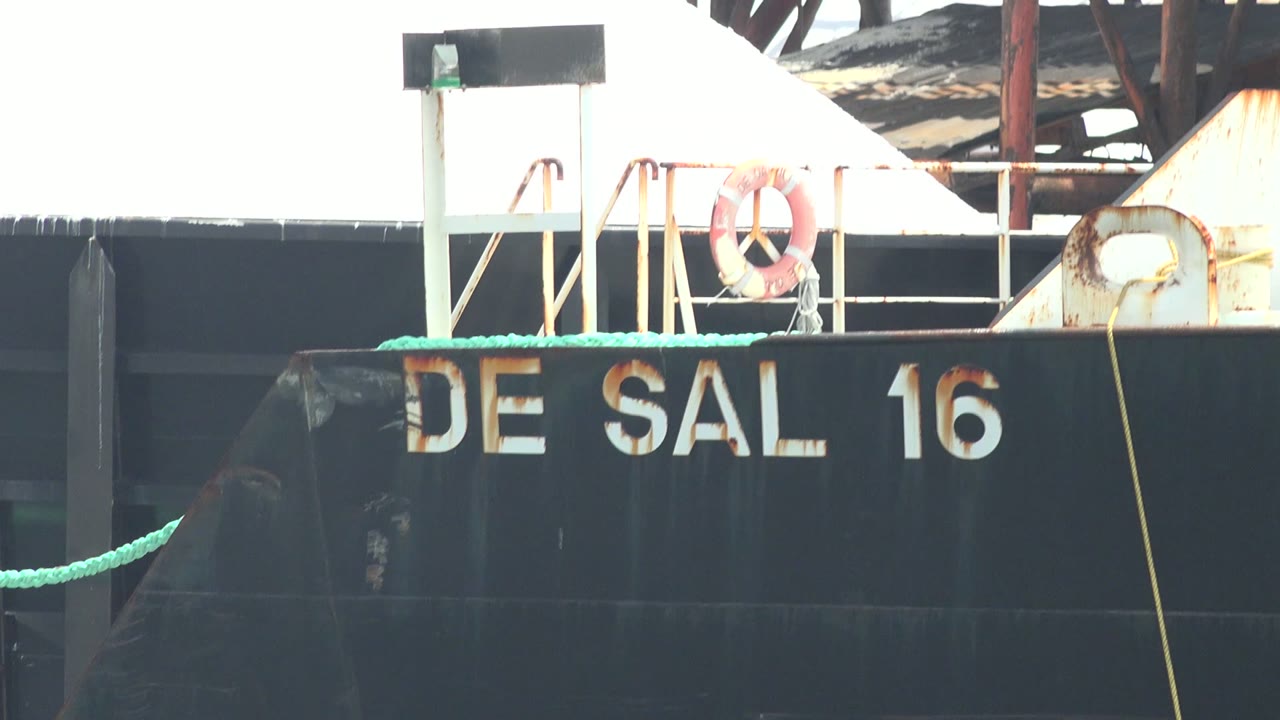A black DE SAL 16 barge in Guerrero Negro, Baja California Sur fills the lower frame, with green rope, a white railing, an orange life ring, rusted metal, and no visible people.
