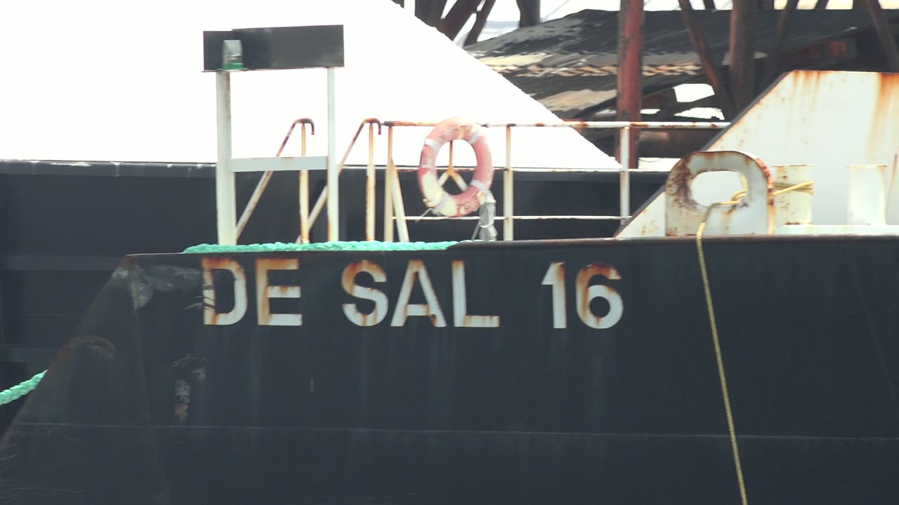 A closer view of the DE SAL 16 barge in Guerrero Negro shows the orange life ring above the black hull, a rusted white metal section to the right, green rope, and no visible people.