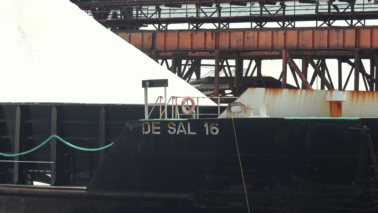 At Guerrero Negro, a black barge marked DE SAL 16 sits close to frame with a mound of white cargo and rusted industrial structures behind it, with no visible people.