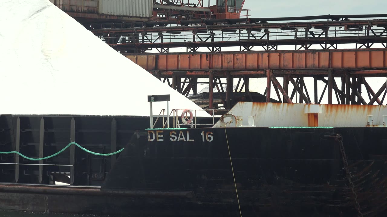 White cargo pile on a black barge under conveyor structures.