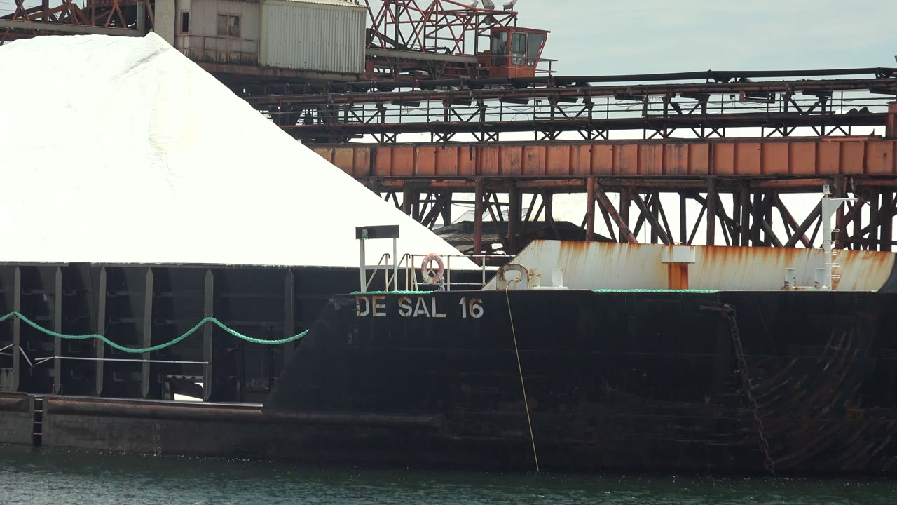 At Guerrero Negro, the bow area of the DE SAL 16 barge appears with green rope along the side, white cargo above, crane trusses behind, and no visible people.