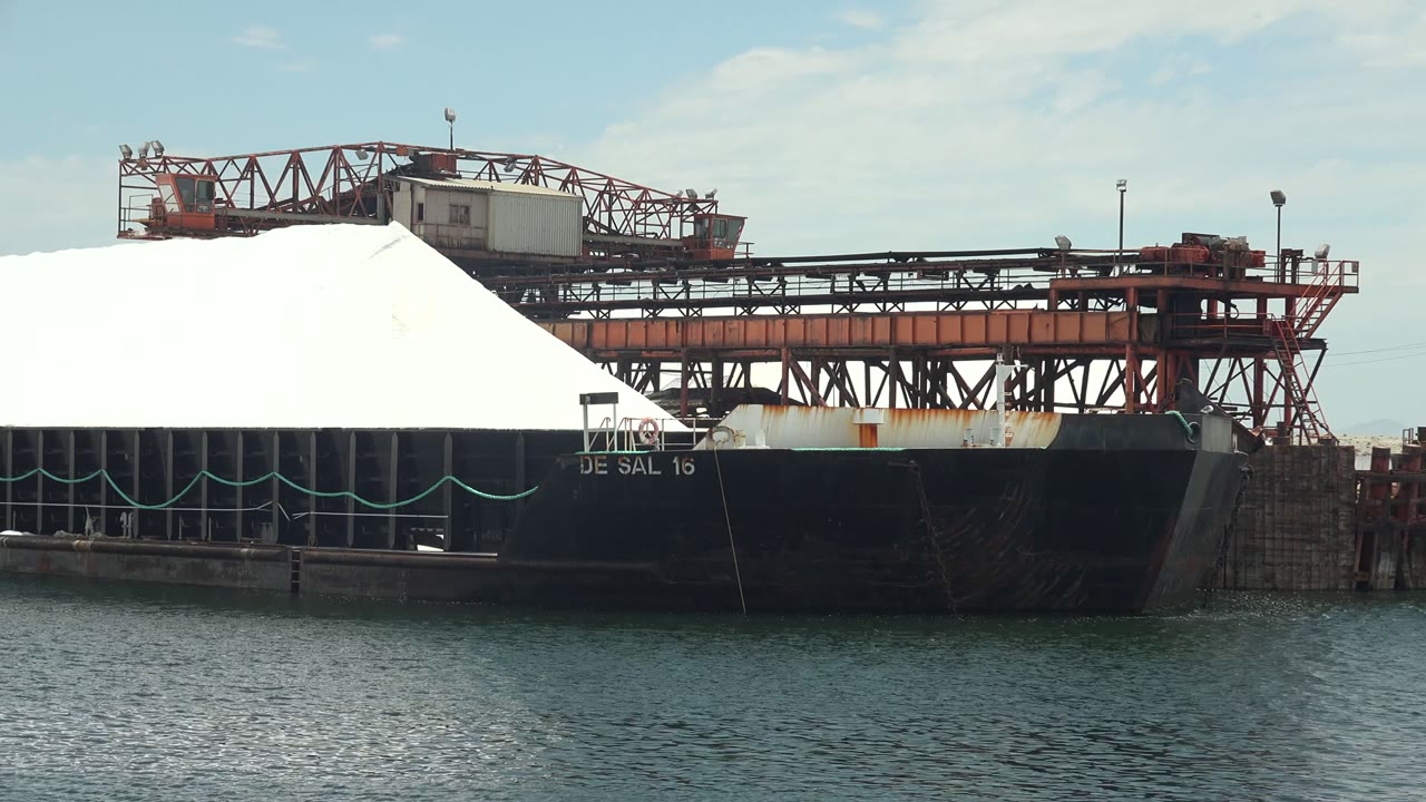 Opening frame in Guerrero Negro, Baja California Sur, showing a black cargo ship beside calm water with a large white cargo mound or cover and red conveyor machinery overhead, no visible people.