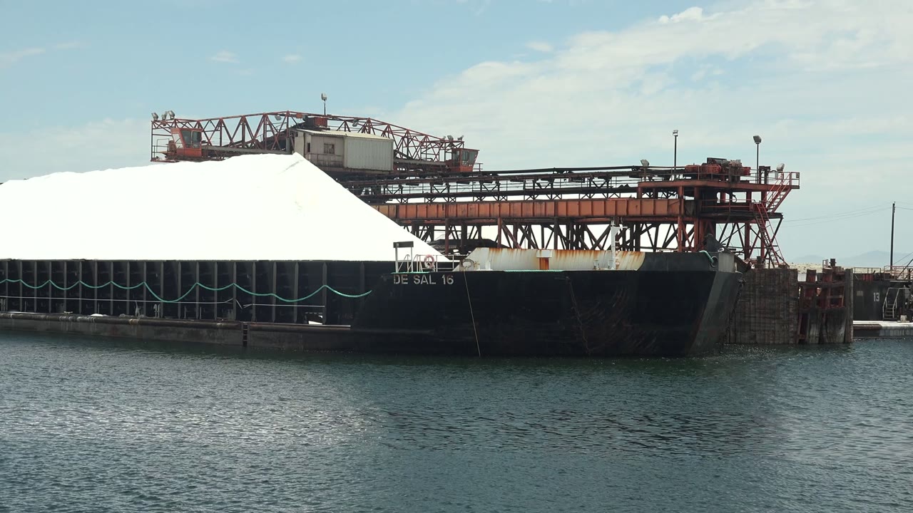 At 0:02, the Guerrero Negro port scene shows more open water in front of the ship while the white cargo surface and red conveyor frame the upper deck, no visible people.