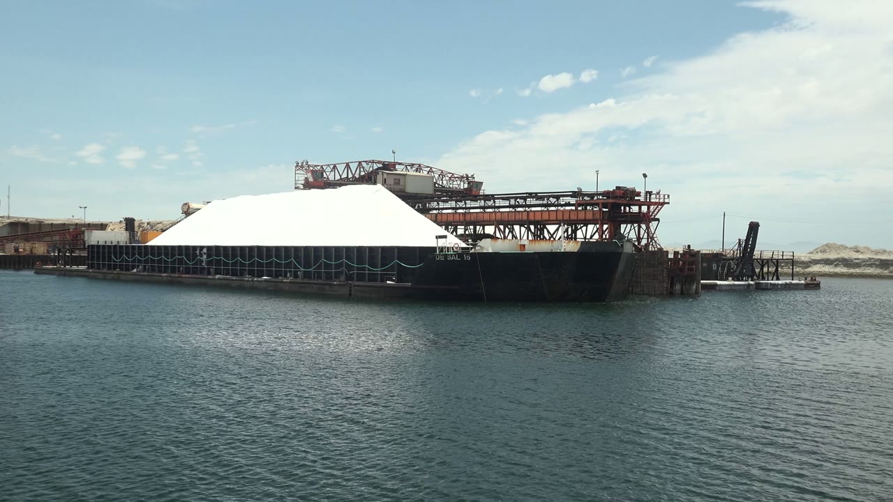 At 0:00 in Guerrero Negro, Baja California Sur, a dark barge with a white dome sits beside an industrial dock across rippled harbor water, with cranes and no visible people.