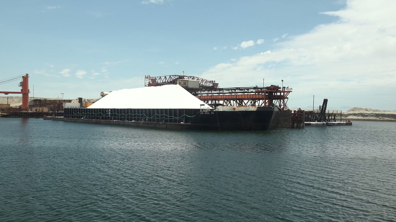 At 0:01, the white dome on the docked barge remains prominent, with red crane equipment behind it and blue-green water filling the foreground in Guerrero Negro, with no visible people.