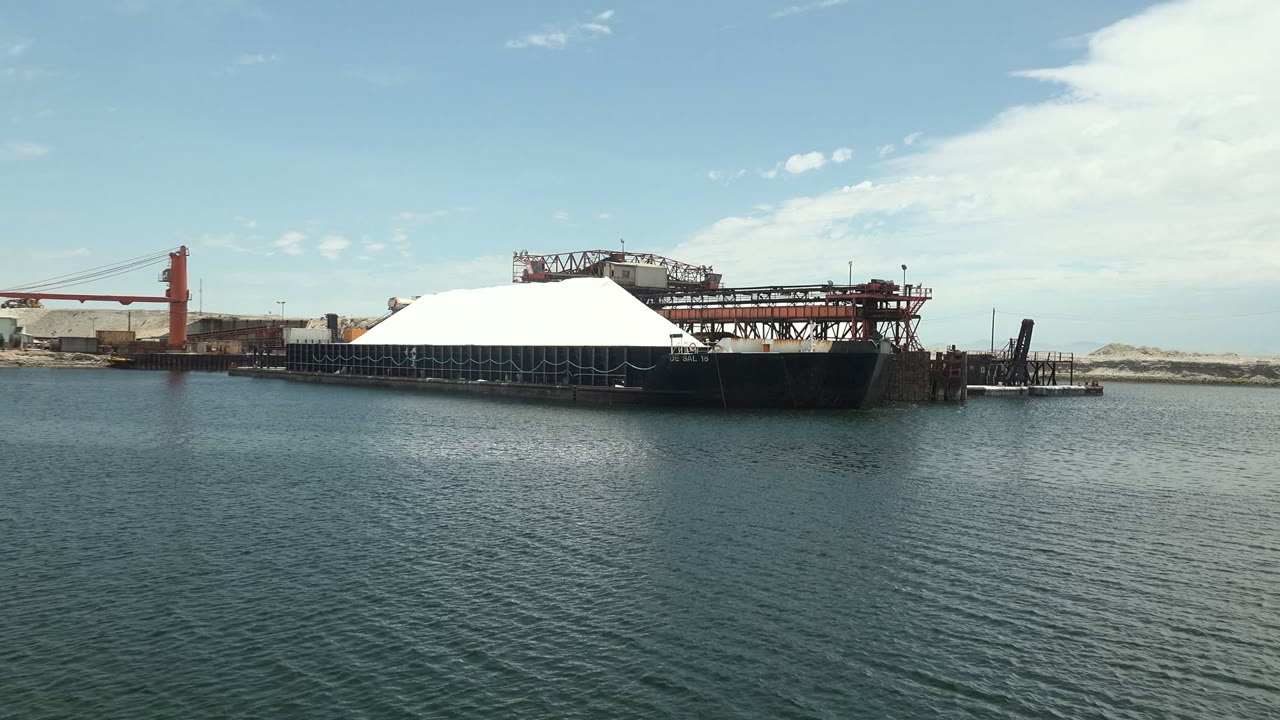 At 0:02 in Guerrero Negro, the barge sits slightly right of center with the white dome, cranes, dock pilings, pale sand flats, open sky, and no visible people.