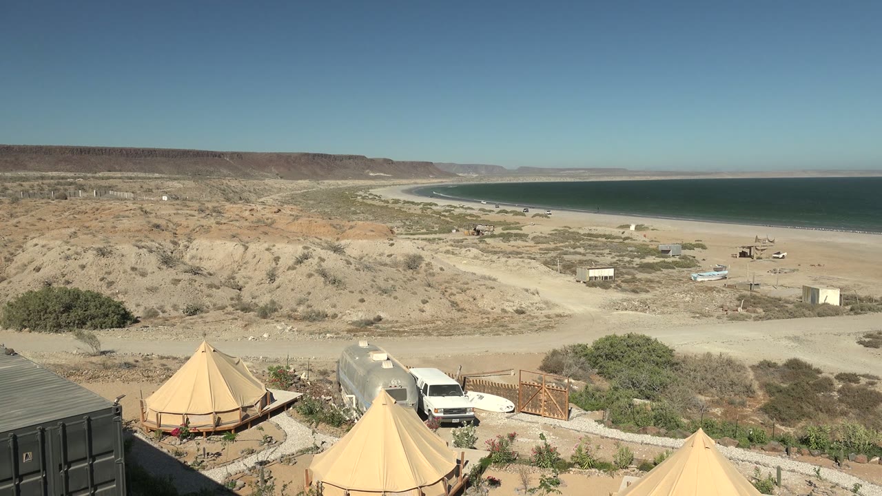 Wide view over a San Juanico campsite with tan tents, a white van, sandy beach, blue sea, arid hills, clear sky, and no visible people.
