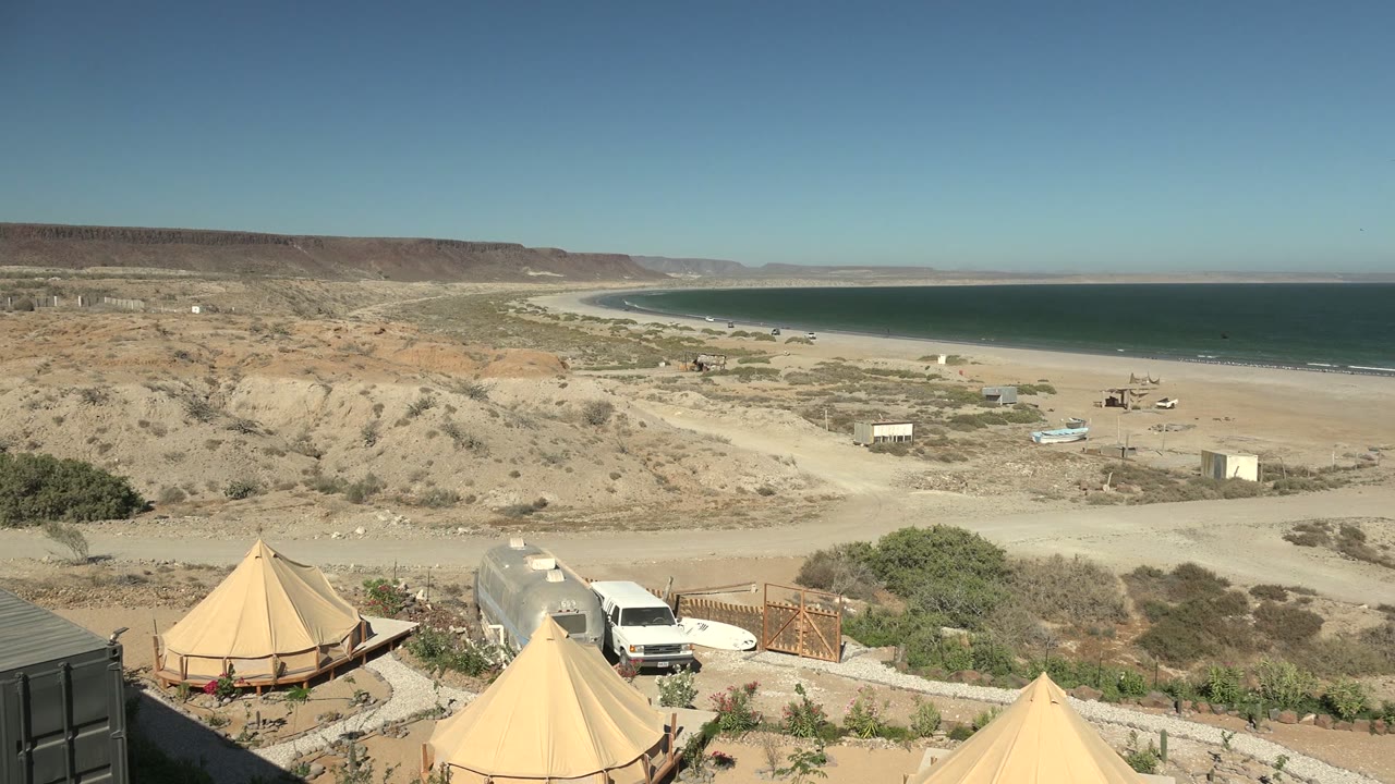 Rightward pan moment in San Juanico with the tents and white van still low in frame, scrubland and beach in the middle distance, and no visible people.