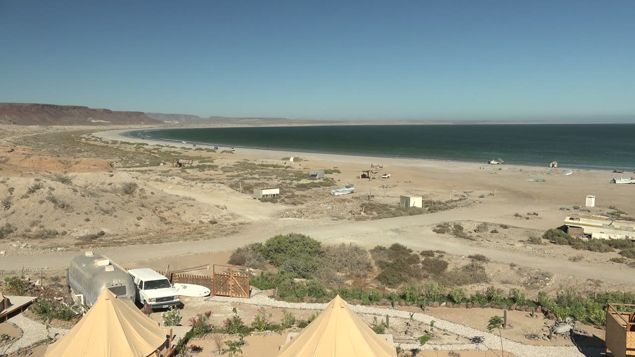 Wide starting view of San Juanico, Baja California Sur with beige tents at the bottom edge, a white van near a gate, sandy beach, blue ocean, distant cliffs, and no visible people.