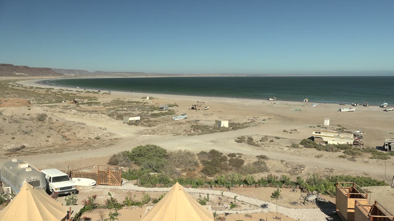San Juanico coastal desert frame with two beige tent peaks low in view, a pale beach road, scattered small buildings, parked vehicles, blue water, and no visible people.