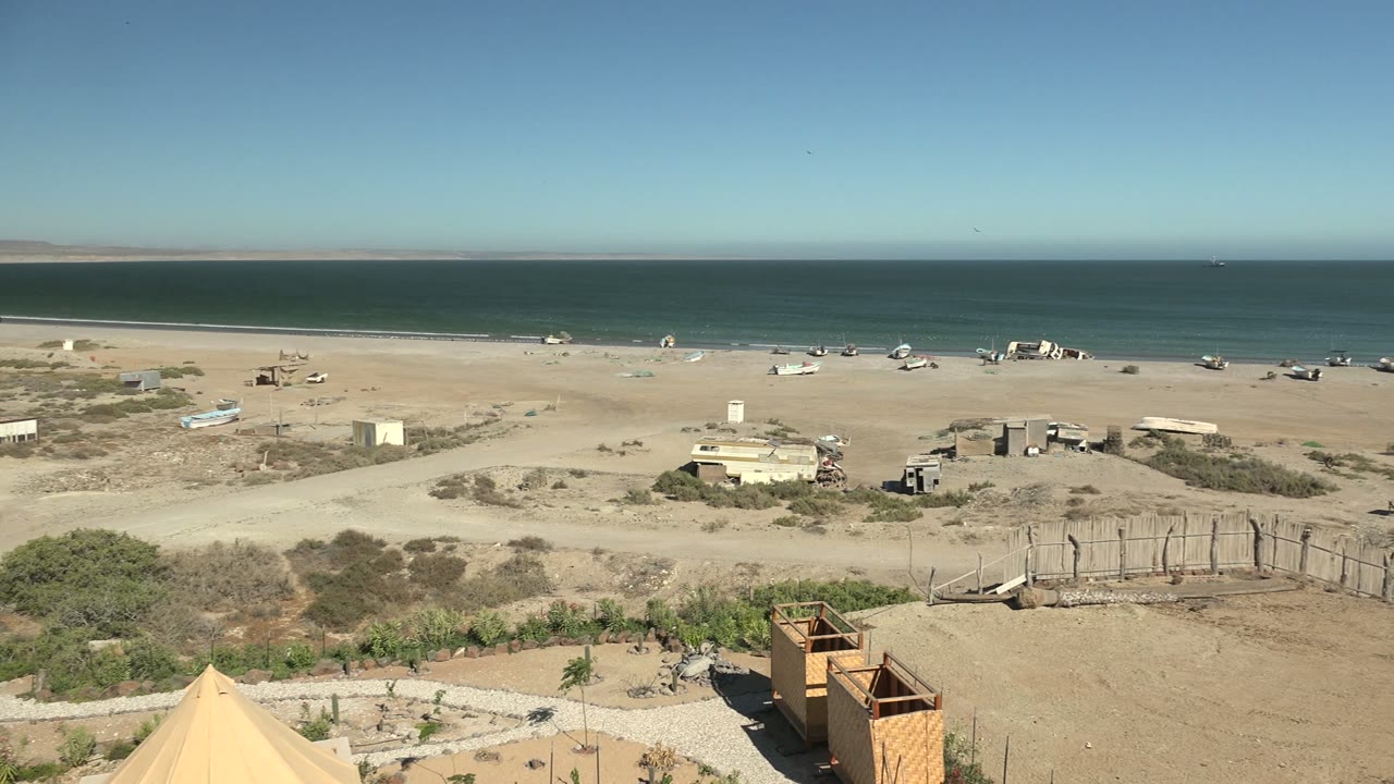 Wide view of San Juanico beach in Baja California Sur with a foreground tent edge, sandy lots, rough fencing, scattered structures, blue ocean, clear sky, and no visible people.