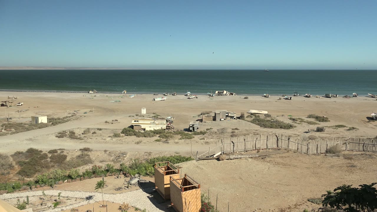 Wide San Juanico beach frame with a high ocean horizon, pale sand, scattered structures near the shore, fenced sandy lots in the midground, and no visible people.