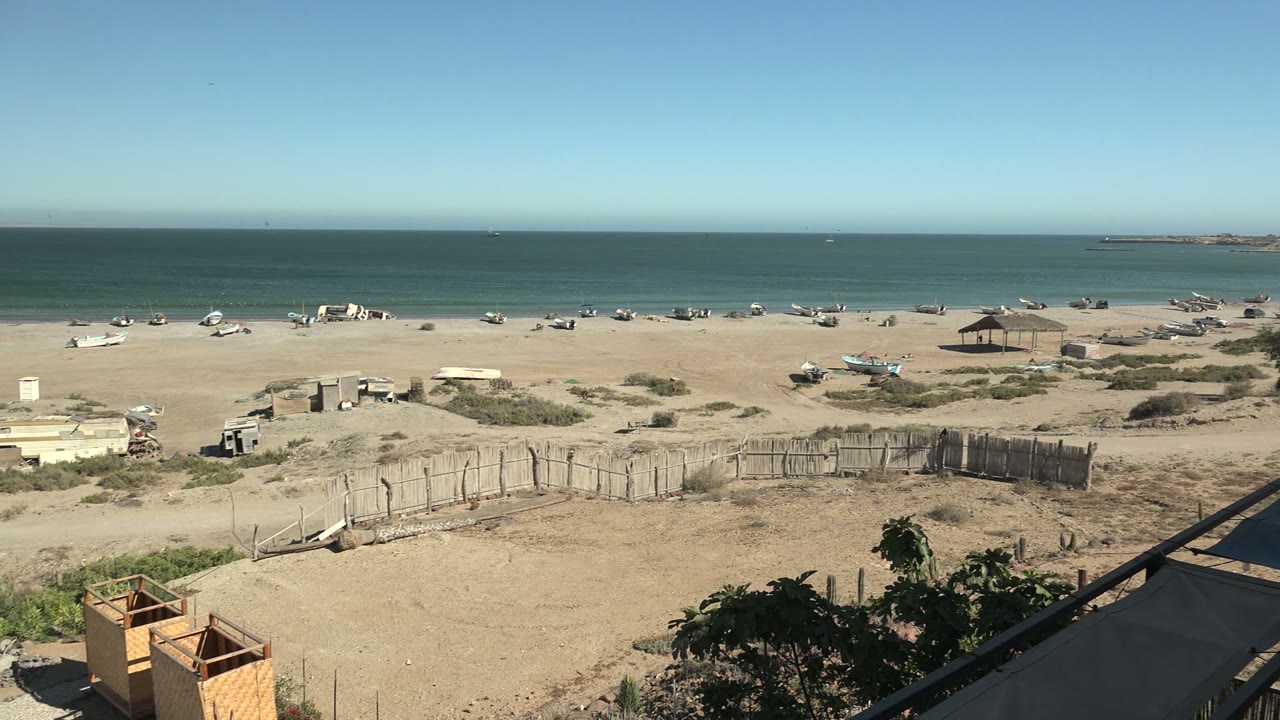 Opening frame of San Juanico beach in Baja California Sur with small boats scattered on the sand, a fence in the foreground, blue ocean beyond, and no visible people.