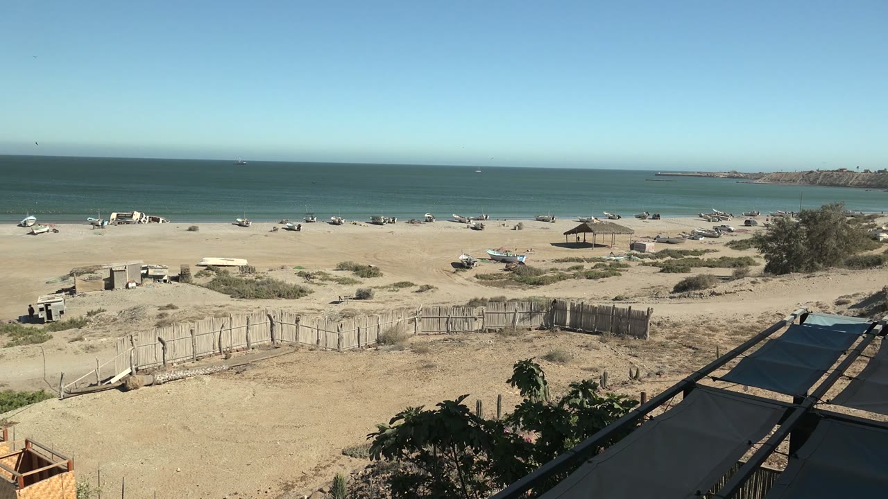 San Juanico beach panorama with boats along the shore, dune scrub, a clear ocean horizon, a rocky point to the right, and no visible people.