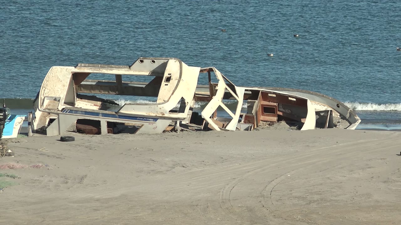Wrecked white boat hull rests partly buried in sand beside blue water in San Juanico, Baja California Sur, with a tire nearby and no visible people.