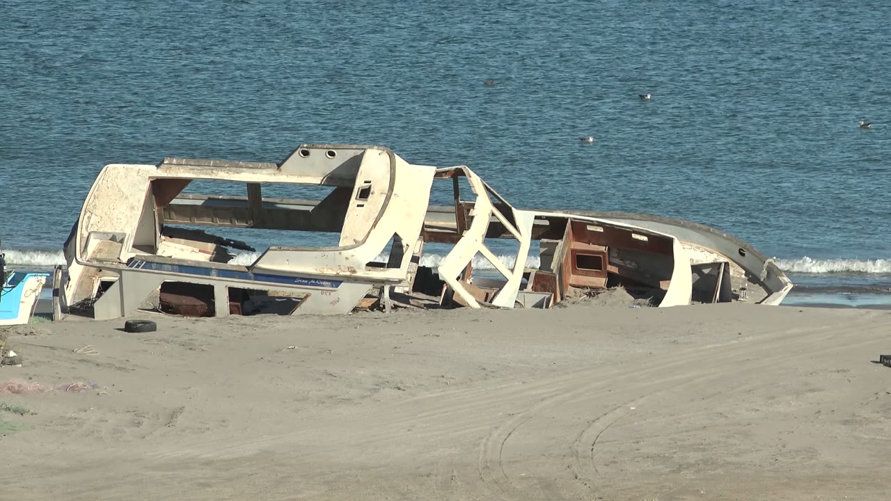Broken boat hull on sand with ocean waves behind it.