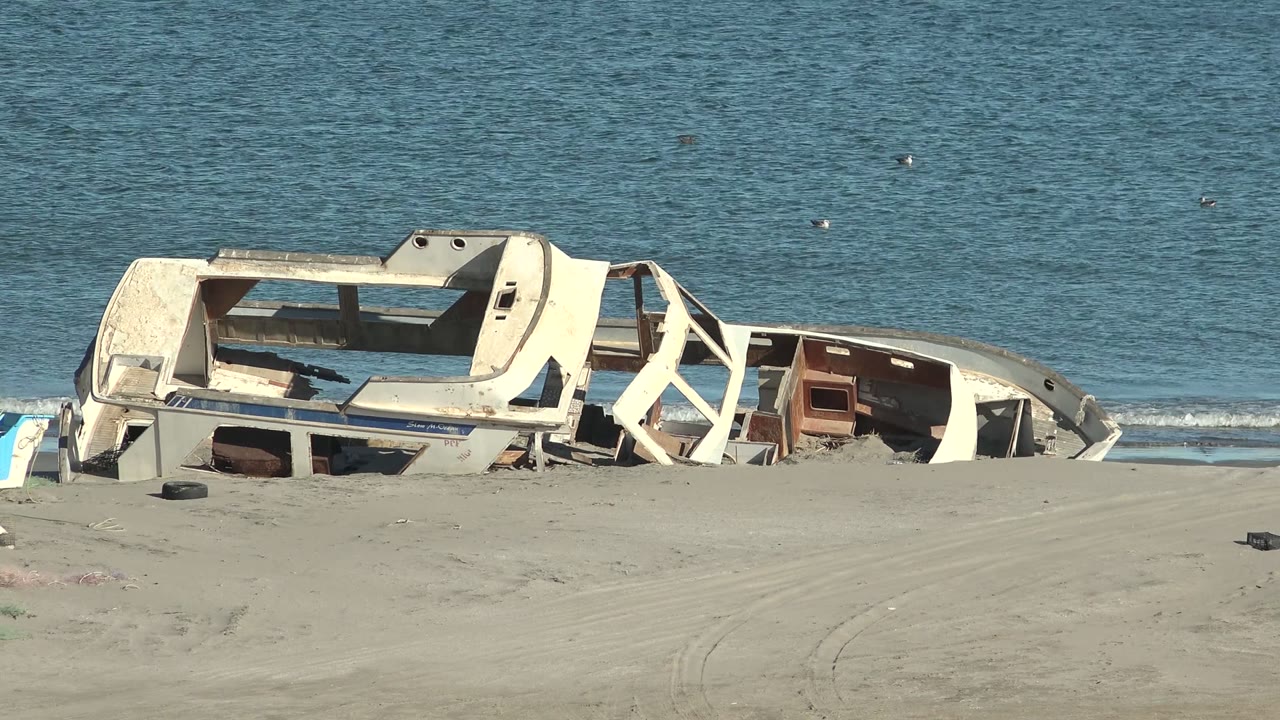 Abandoned white boat wreck fills the midground on the San Juanico beach, with beige sand in front, calm blue water behind, and no visible people.
