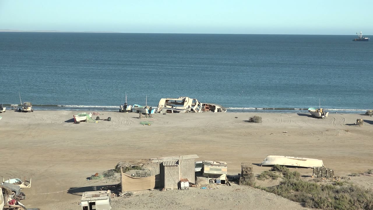 Wide view of San Juanico beach in Baja California Sur with small boats on pale sand, calm blue ocean, distant offshore boats, low foreground structures, and no visible people.