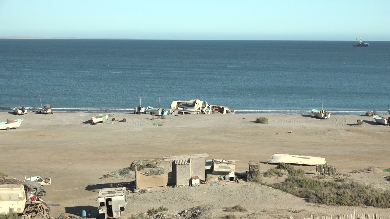 Sandy shoreline with several small boats, blue ocean, clear horizon, and simple beach structures.