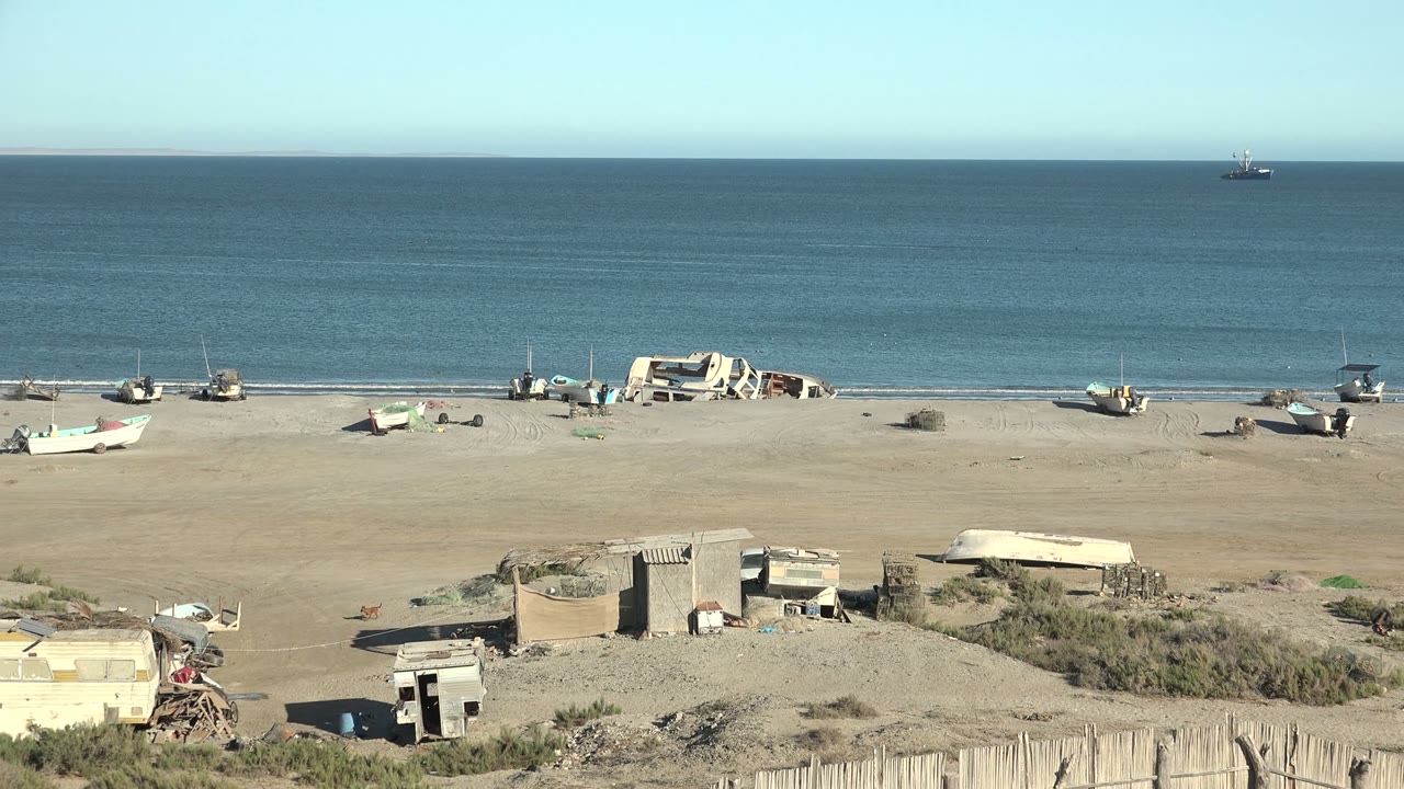 Wide San Juanico beach view with boats near the waterline, calm sea under pale blue sky, rough foreground shelters, scrub patches, and no visible people.