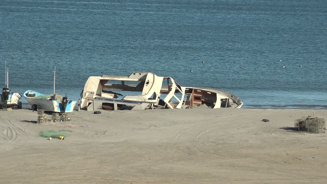 At 0:00 in San Juanico, Baja California Sur, a wrecked white boat hull sits near small boats on a sandy beach with calm blue ocean behind it and no visible people.