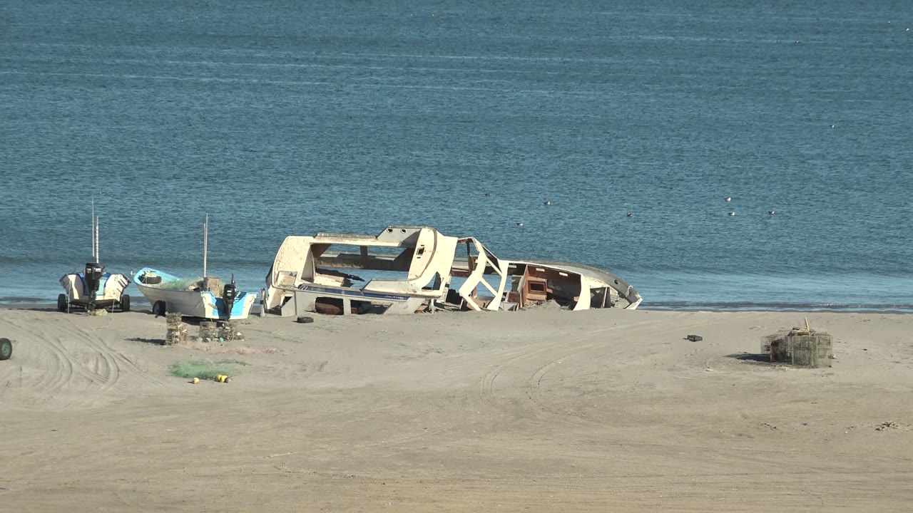 Wrecked boat hull, beach debris, and calm water from a similar shoreline view.